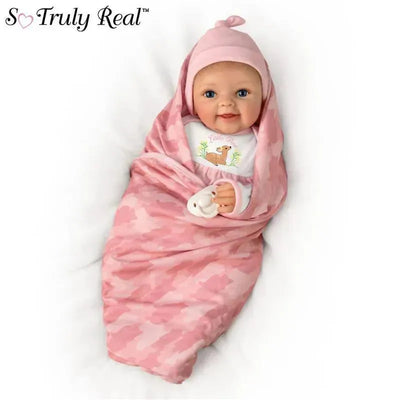 Lifelike baby girl doll wrapped in a pink camouflage blanket, wearing a matching hat and a shirt featuring a fawn illustration.