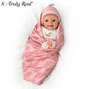 Lifelike baby girl doll wrapped in a pink camouflage blanket, wearing a matching hat and a shirt featuring a fawn illustration.