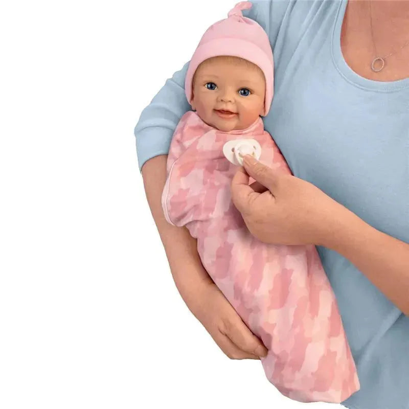 Lifelike baby girl doll wrapped in a pink camouflage blanket, held by a person wearing a light blue top, smiling and holding a pacifier.