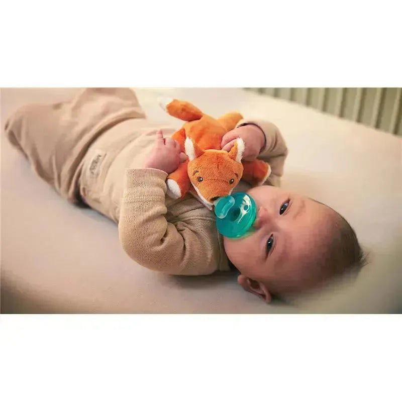 Infant lying on a blanket, cozily holding a plush orange fox toy and using a teal pacifier.