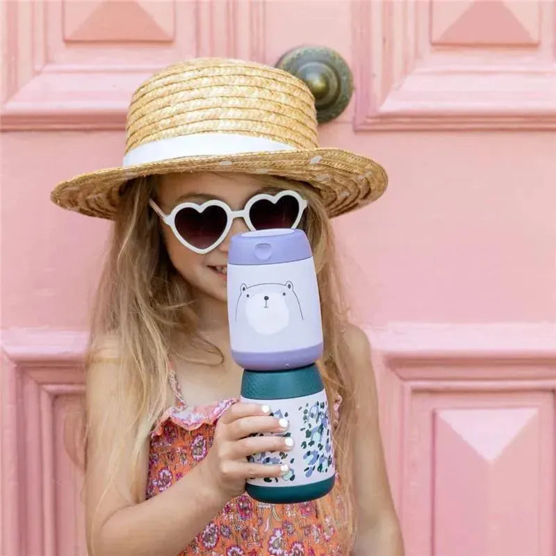 Child wearing a straw hat and heart-shaped sunglasses holds a B.box Bear Hugs Insulated Food Jar Mini in front of a pink door.