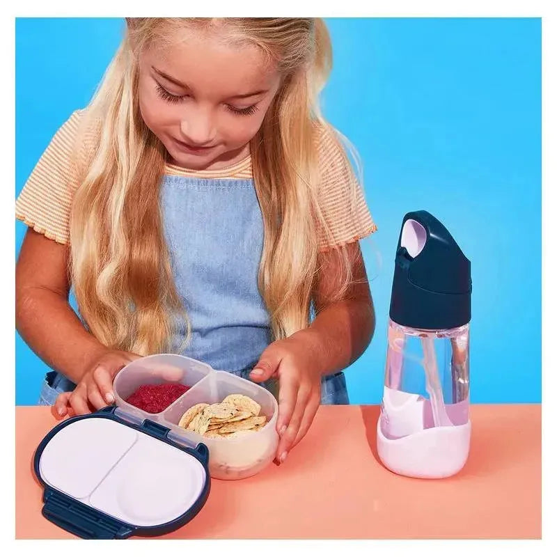 A child with long hair examines a B.box Snackbox containing snacks, alongside a clear water bottle.