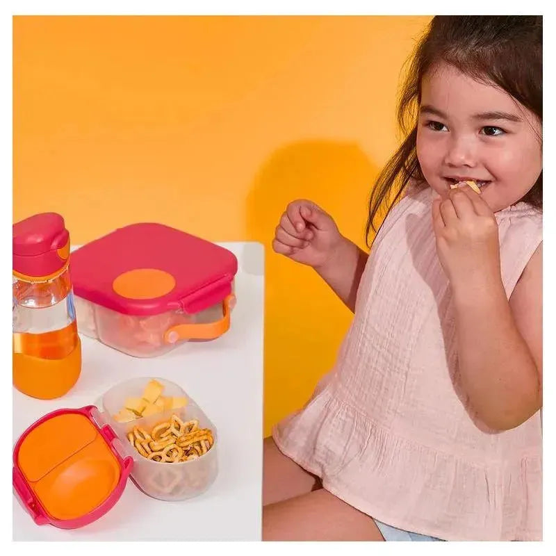 A young girl is enjoying a snack while sitting at a table with a pink B.box Snackbox containing pretzels and cheese. A water bottle is nearby.