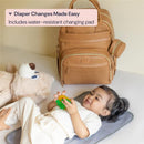 Light brown vegan leather diaper bag beside a toddler lying on a changing pad, playing with a toy.