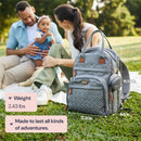 Travel essentials baby bag in brown with a changing pad sits on grass, beside a couple enjoying time with their baby.