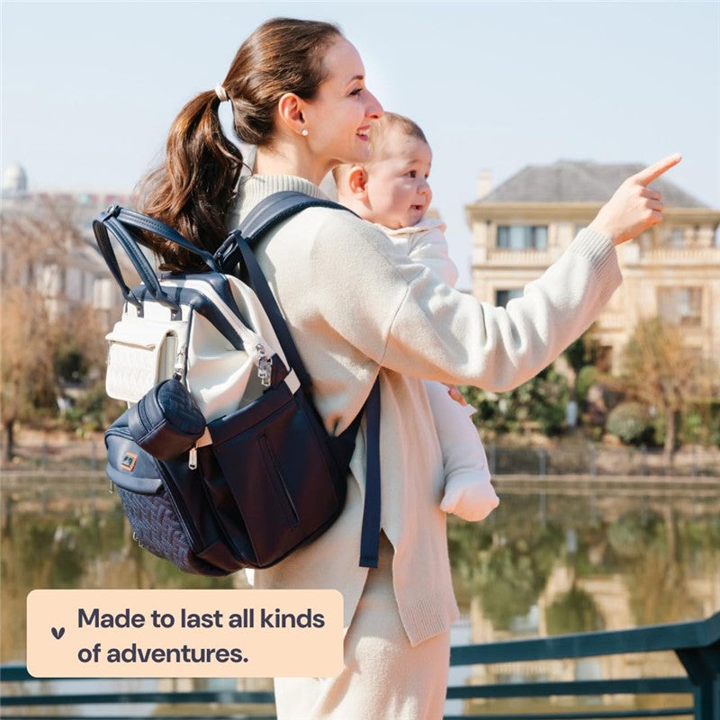 A woman holds a baby while wearing a BabbleRoo vegan leather diaper bag backpack in blueberry cream.