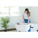 A woman gently bathes a baby in a white Babu Bath luxury bathtub with a wooden stand and a blue towel, near a window.