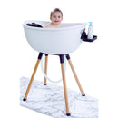 Luxury white baby bathtub on a wooden stand, with a smiling baby inside and a bottle of soap on the side.