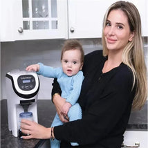 Mom holds a baby while using the Baby Brezza Formula Pro Mini to prepare formula in a kitchen setting.