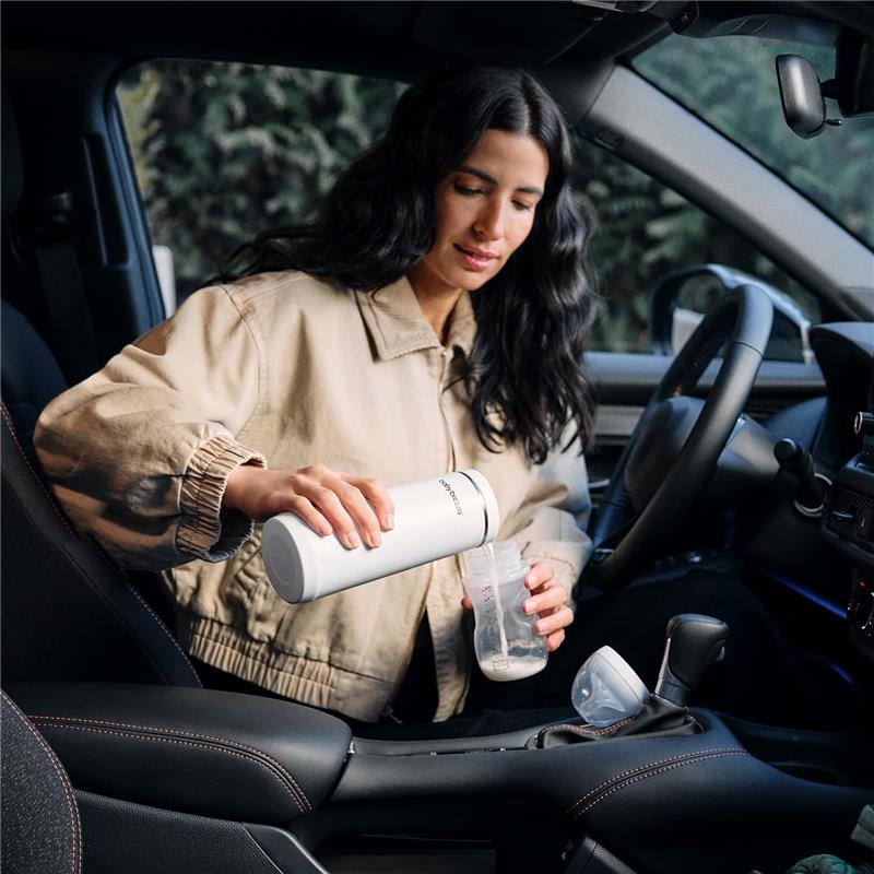 A woman with long dark hair pours milk from a Baby Brezza portable bottle warmer into a baby bottle while sitting in a car.