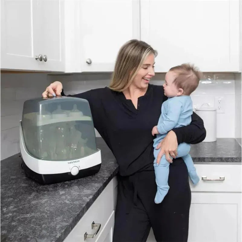 A woman holds a baby while interacting with a Baby Brezza Superfast Sterilizer Dryer on a kitchen countertop.