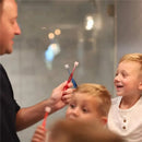 A man holds two toothbrushes, one red and one blue, while smiling children look on, engaging in a fun, educational moment.