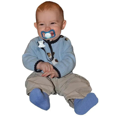 A smiling baby wearing a light blue sweater and beige pants holds a pacifier, showcasing the Baby Buddy pacifier holder in pink.