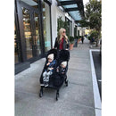 Double stroller with two children inside, pushed by a woman in a black leather jacket sunglasses; outdoor setting.