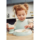 Child wearing a Babybjorn powder green bib, exploring food with a spoon and drinking from a matching cup.