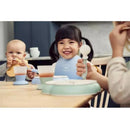 Two children at a dining table with Babybjorn bibs; one smiles with a spoon, the other eats a snack.