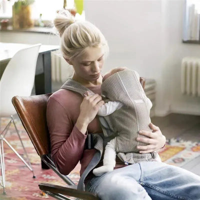 Woman sitting in a chair, using a Babybjorn Baby Carrier Mini in gray beige to hold an infant close.