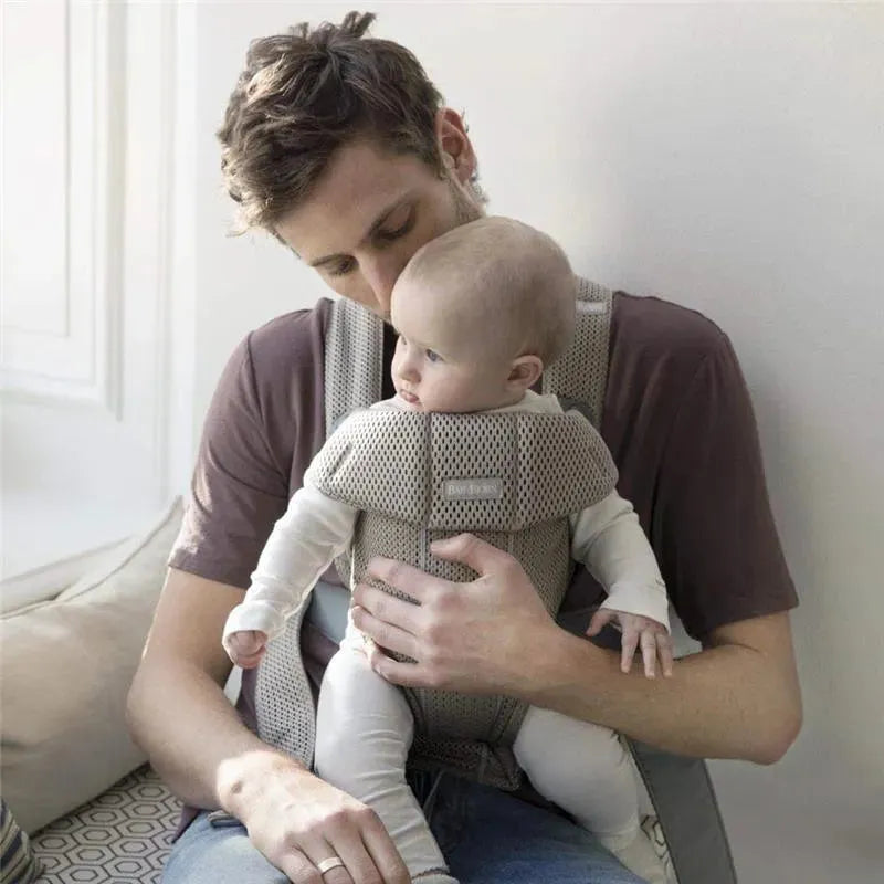 Babybjorn Baby Carrier Mini in gray beige, supporting an adult holding a baby, seated on a couch, with a soft background.