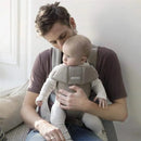 Babybjorn Baby Carrier Mini in gray beige, supporting an adult holding a baby, seated on a couch, with a soft background.