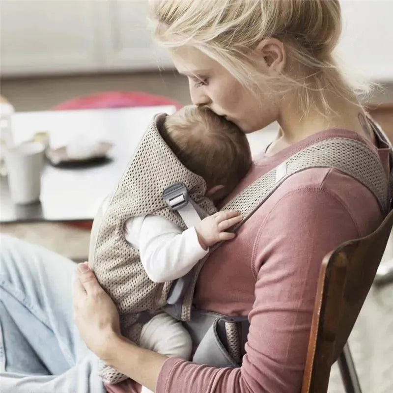 Mother gently kisses her baby in a Babybjorn Baby Carrier Mini, made of gray beige 3D mesh, while sitting in a chair.