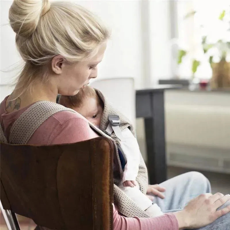 A woman sits in a chair, gently cradling her baby in a gray-beige Babybjorn carrier made of 3D mesh.