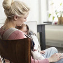 A woman sits in a chair, gently cradling her baby in a gray-beige Babybjorn carrier made of 3D mesh.