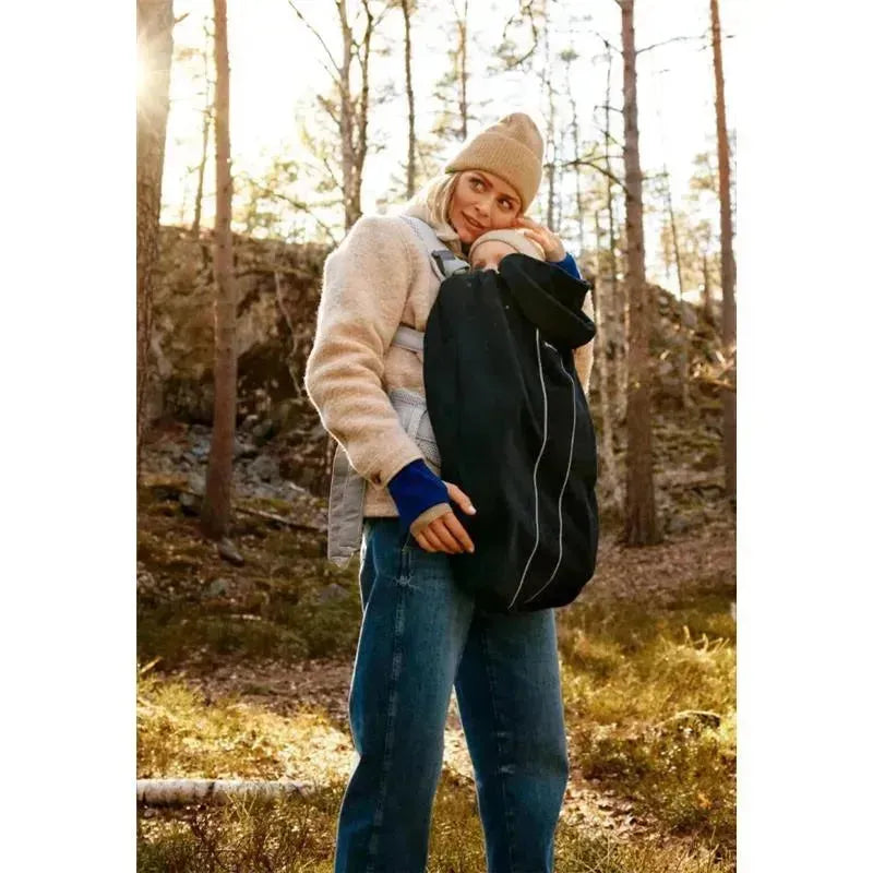 Mother holding a baby in a black Babybjorn carrier cover while standing in a forest setting.