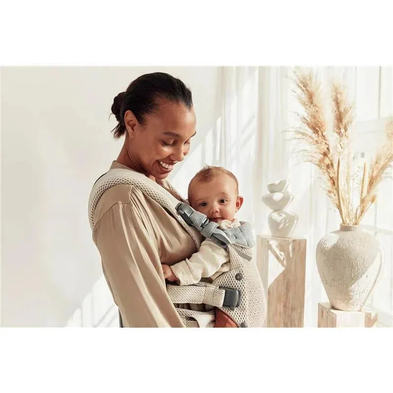 A smiling woman holds a baby in a BabyBjorn Carrier Harmony 3D Mesh, Cream, while surrounded by a bright, airy room.