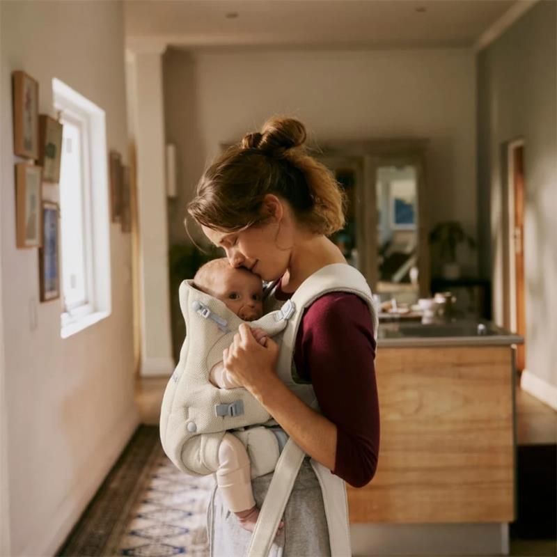 Mother gently kisses her baby in a Babybjorn Baby Carrier Hold Mesh, cream, in a warmly lit home environment.