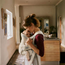 Mother gently kisses her baby in a Babybjorn Baby Carrier Hold Mesh, cream, in a warmly lit home environment.