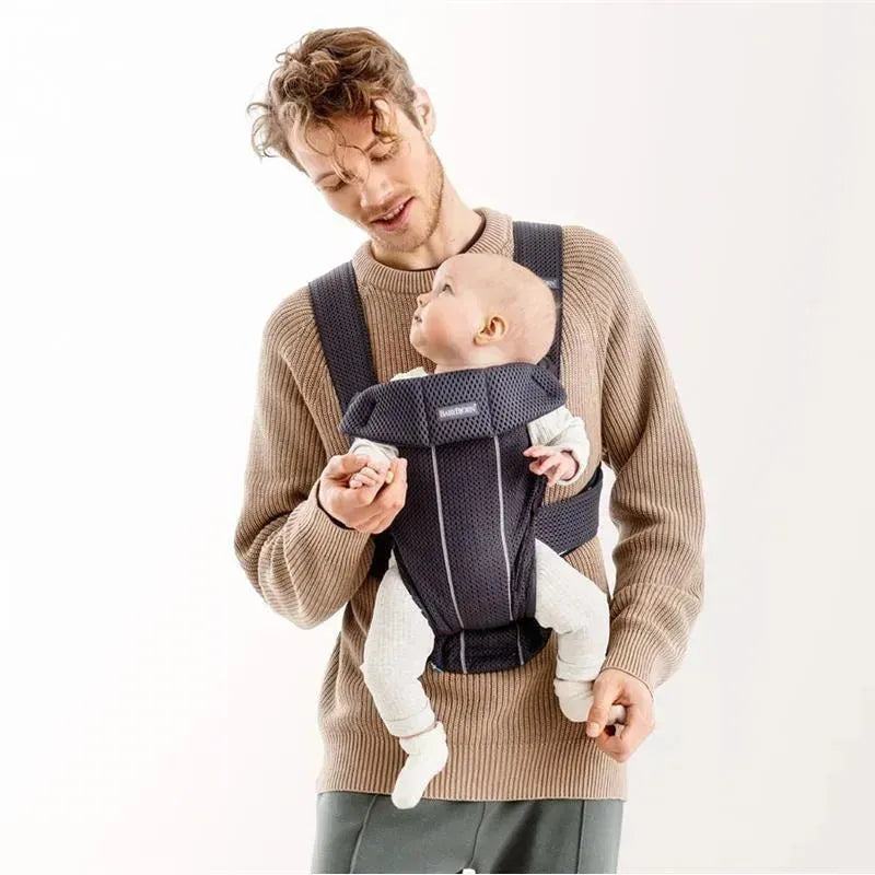 A man wearing a beige sweater carries an infant in a Babybjorn Baby Carrier Mini 3D Mesh, Anthracite, smiling at the baby.
