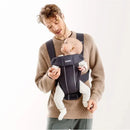 A man wearing a beige sweater carries an infant in a Babybjorn Baby Carrier Mini 3D Mesh, Anthracite, smiling at the baby.