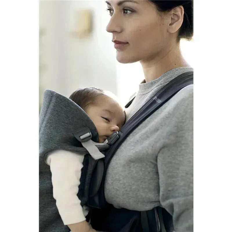 A mother wearing a BabyBjorn Baby Carrier Mini in Dark Gray, holding a sleeping baby close to her chest.