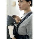 A mother wearing a BabyBjorn Baby Carrier Mini in Dark Gray, holding a sleeping baby close to her chest.