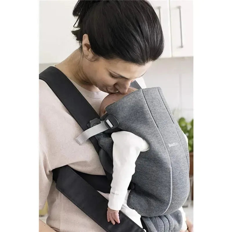 BabyBjorn Baby Carrier Mini in dark gray, featuring a caregiver tenderly kissing a baby securely positioned inside.