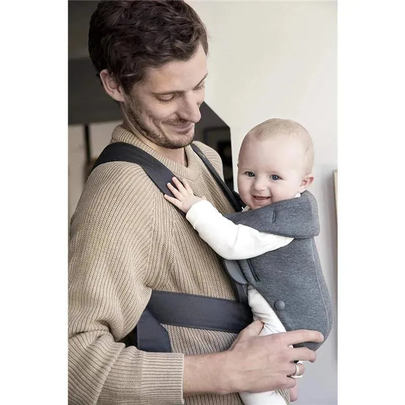 BabyBjorn Baby Carrier Mini in dark gray is worn by a smiling father holding a happy baby in a cozy indoor setting.