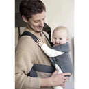 BabyBjorn Baby Carrier Mini in dark gray is worn by a smiling father holding a happy baby in a cozy indoor setting.