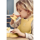Child in a yellow bib sipping from a pink cup, with a baby dinner set visible on the table.