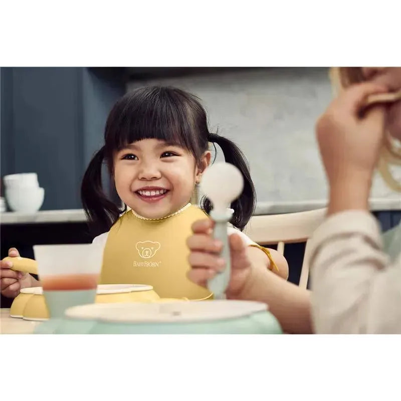 Smiling child wearing a yellow BabyBjorn bib, holding a spoon, sitting at a table with a powder blue dinner set.