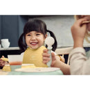 Smiling child wearing a yellow BabyBjorn bib, holding a spoon, sitting at a table with a powder blue dinner set.
