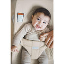 BabyBjörn Bouncer Balance Soft in light beige with a smiling baby seated, held by an adult, displaying safety label.