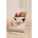 Light beige BabyBjörn Bouncer Balance Soft with a baby sitting, holding a small card, on a textured rug.