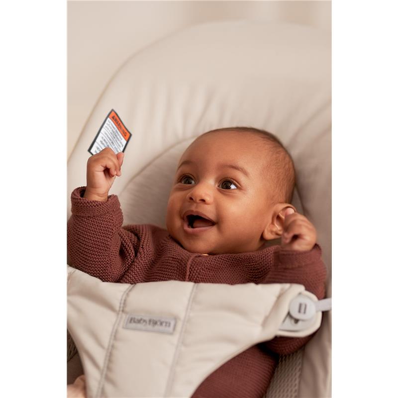 Infant joyfully sitting in a BabyBjörn Bouncer Balance Soft, light beige, wearing a brown sweater.