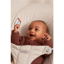 Infant joyfully sitting in a BabyBjörn Bouncer Balance Soft, light beige, wearing a brown sweater.