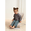 Child sitting in a BabyBjörn Bouncer Balance Soft in light beige, smiling, wearing a gray sweater and blue pants.