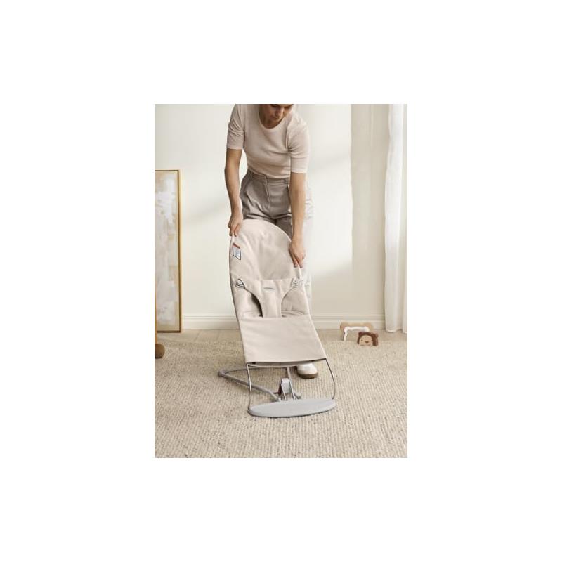 A woman adjusts the Babybjorn Bouncer Bliss in Light Beige, showcasing its fabric and ergonomic design in a home setting.