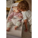 Light beige Babybjorn Bouncer Bliss with a smiling baby in a pink sweater and a child leaning in to interact.