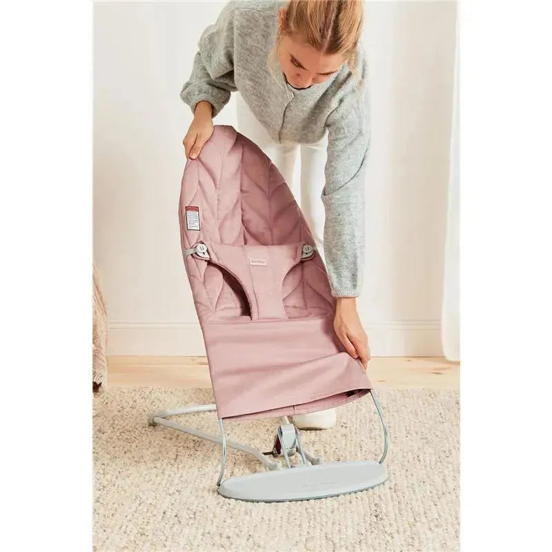 A person adjusts the Babybjorn Bouncer Bliss in dusty pink, featuring a quilted seat and a lightweight frame.