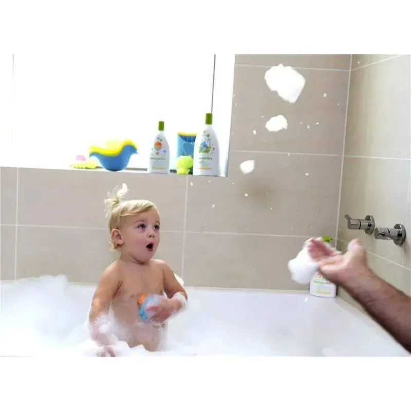 A surprised toddler in a bubble bath plays with bath toys while bubbles float around. Babyganics products are visible in the background.