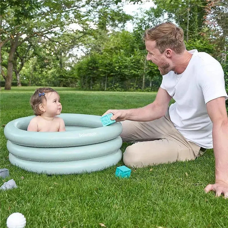 Inflatable Babymoov baby bathtub in a grassy area with a smiling parent playing with a child inside.